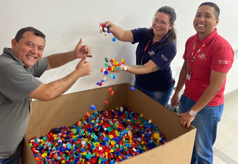 Foto de voluntários próximos a uma caixa de papelão com milhares de tampinhas plásticas.