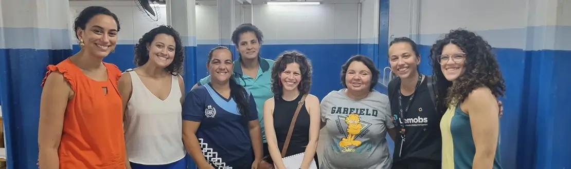 Foto de uma menina de costas sentada em uma cadeira em uma sala de aula. À sua esquerda, há a mão de uma pessoa que segura um tablet. A menina está interagindo com a pesquisa que está na tela do equipamento.