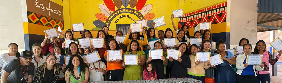 Foto de um grupo diverso de pessoas, em pé e bem juntinhas, em frente a uma parede pintada com temática indígena da Escola Tengatuí Marangatu. Algumas das pessoas exibem nas mãos certificados de participação do curso.