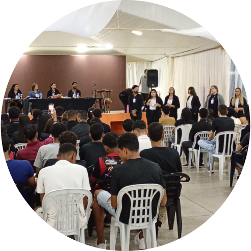 Foto de um auditório onde estudantes estão sentados em cadeiras voltadas para a frente, assistindo a uma apresentação conduzida por um grupo de pessoas posicionadas ao lado de um palco, enquanto uma mesa com outras pessoas compõe a banca ao fundo.