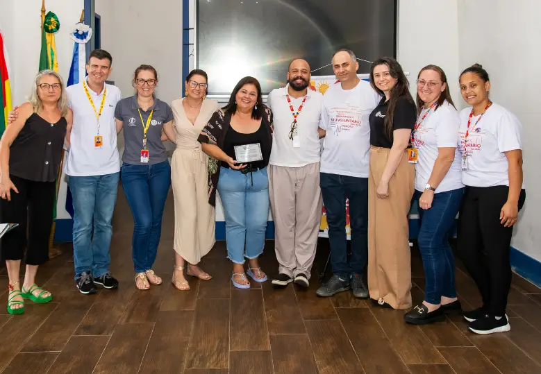 Foto de um grupo de pessoas em pé no interior de uma sala. Uma delas segura uma placa de metal nas mãos. Todas sorriem.