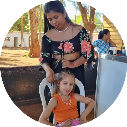 Uma mulher indígena fazendo tranças no cabelo de uma menina criança da comunidade. No fundo, há uma mulher olhando para o lado.