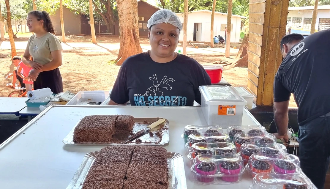 Midian, que produz bolos e doces para venda na comunidade, sorrindo para a câmera e exibindo pedaços de bolo e cupcakes de chocolate. No fundo, há adultos e crianças.