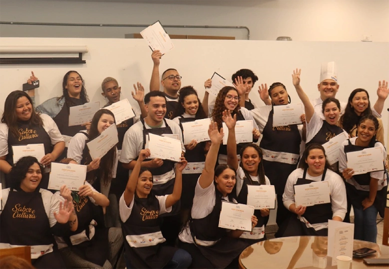 Foto do grupo de estudantes e do chef instrutor do curso, todos juntos, alguns em pé e outros abaixados, segurando seus certificados nas mãos.