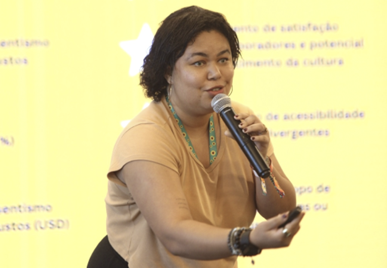 Foto de Sarah Fernn com um microfone na mão durante sua apresentação no Demoday Iniciativa Potências do Nossa Parte Pela Educação. 
