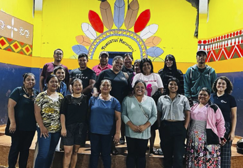 Foto de um grupo de mulheres e homens, dentre eles Jennifer França, do Instituto BRF, em pé e bem próximos uns dos outros. Na parede atrás deles, há o desenho de um cocar e o nome da escola Tengatuí Marangatú.