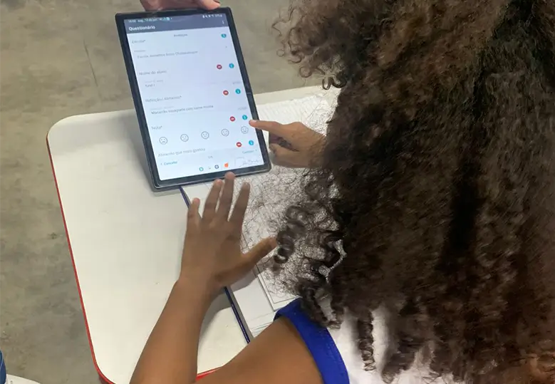 Foto de uma menina de costas sentada em uma cadeira em uma sala de aula. À sua esquerda, há a mão de uma pessoa que segura um tablet. A menina está interagindo com a pesquisa que está na tela do equipamento.