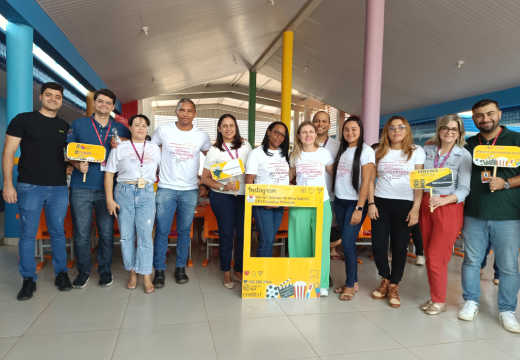 Foto de um grupo de onde pessoas em pé e bem juntinhas em uma escola. Algumas delas vestem camisetas com a nuvem de palavras sobre voluntariado do Instituto BRF e seguram nas mãos placas indicativas da ação Vai ter cinema no meu bairro.