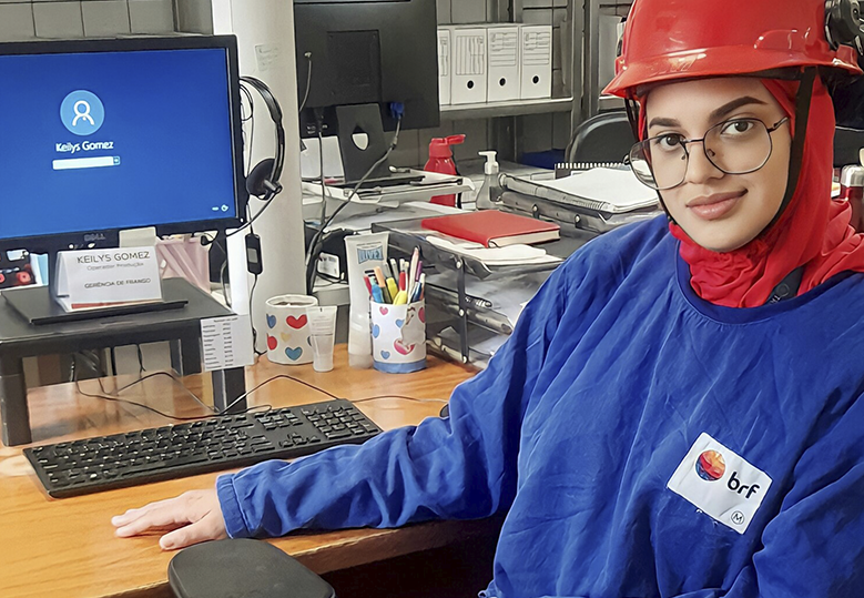 Foto de Keilys Gomes sentada em frente a uma estação de trabalho com computador. Ela usa óculos de grau, está com capacete de proteção na cabeça, usa uniforme azul com logo da BRF e sorri levemente.