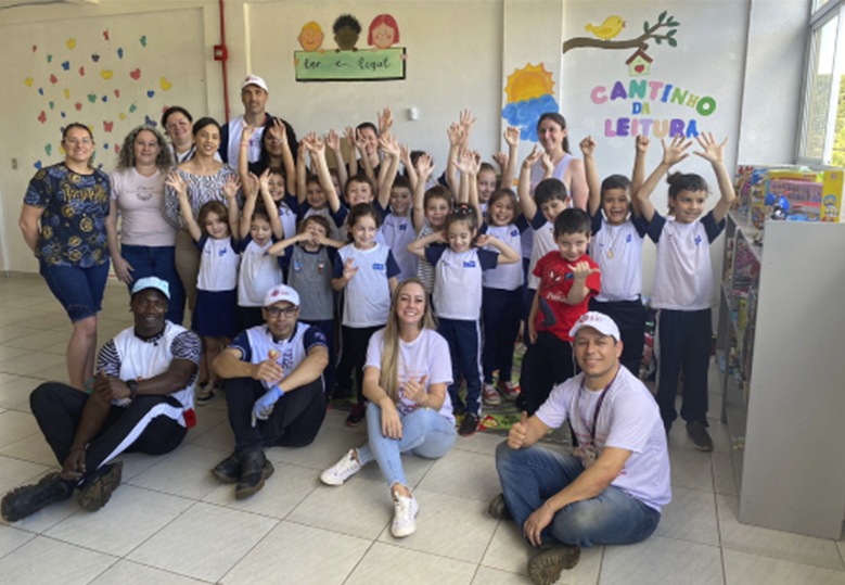 Foto de um grupo de voluntárias e voluntários do Instituto, pessoas adultas e crianças reunidos em uma área interna de uma escola. Alguns estão sentados no chão enquanto outros estão de pé. Todos sorriem.