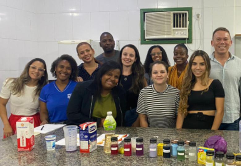Foto de um grupo diverso de pessoas em uma cozinha. Na frente deles, há uma mesa com alimentos e vários potes plásticos.