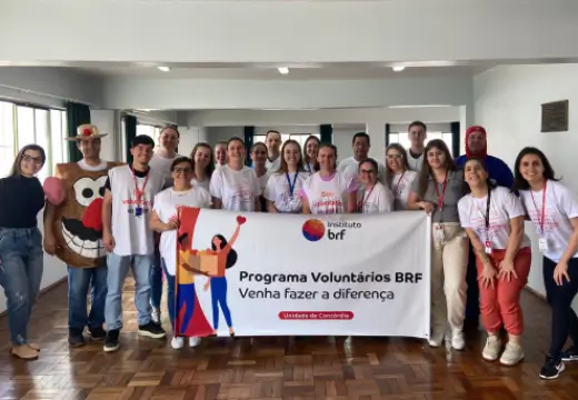 Foto de um grupo diverso de pessoas em uma sala. Elas estão em pé, usam camisetas e coletes do programa de voluntariado do Instituto BRF, e na frente seguram um banner com o logo do Instituto BRF e o texto “Programa Voluntários BRF. Venha fazer a diferença.”