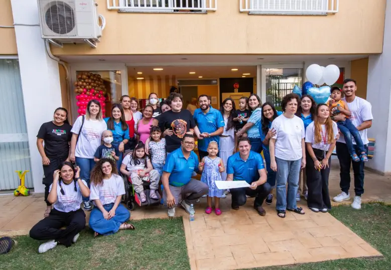 A imagem mostra um grupo diverso de pessoas, incluindo crianças e adultos, reunidos em frente a um edifício decorado com balões. Algumas pessoas usam uniformes azuis e camisetas de voluntariado brancas, e o ambiente transmite um clima alegre e acolhedor.