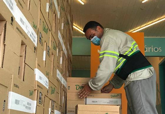 Foto de homem uniformizado e com máscara de proteção cobrindo a boca e o nariz empilhando caixas de produtos Deline.