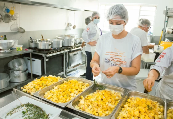 Foto de um grupo de voluntárias do Instituto MBRF e da Gastromotiva em uma cozinha solidária. Todas usam touca para cobrir os cabelos e máscara de proteção para o nariz e a boca. Uma delas está em destaque no primeiro plano, e tira folhas de alecrim e coloca sobre cubas com batatas picadas.