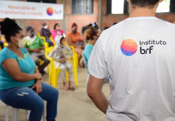 Foto de um homem, em pé, de costas para a câmera. Ele veste uma camiseta branca com o logo do Instituto MBRF. A sua frente, desfocadas, há mulheres e crianças sentadas em cadeiras de plástico.