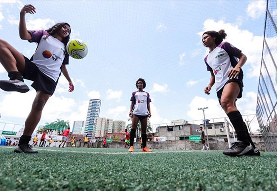 Foto de três meninas em um campo de futebol, formando um triângulo. Uma delas está com a perna levantada pronta para bater a bola que está suspensa no ar. Todas estão uniformizadas, e há o logo do Instituto MBRF em suas camisetas.